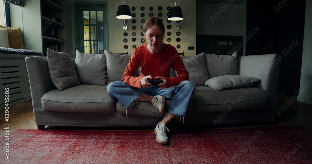 Obraz premium A young woman in casual clothes is smiling while playing video games with a joystick, sitting on the couch at home. The video moves from the general plan to the woman's face