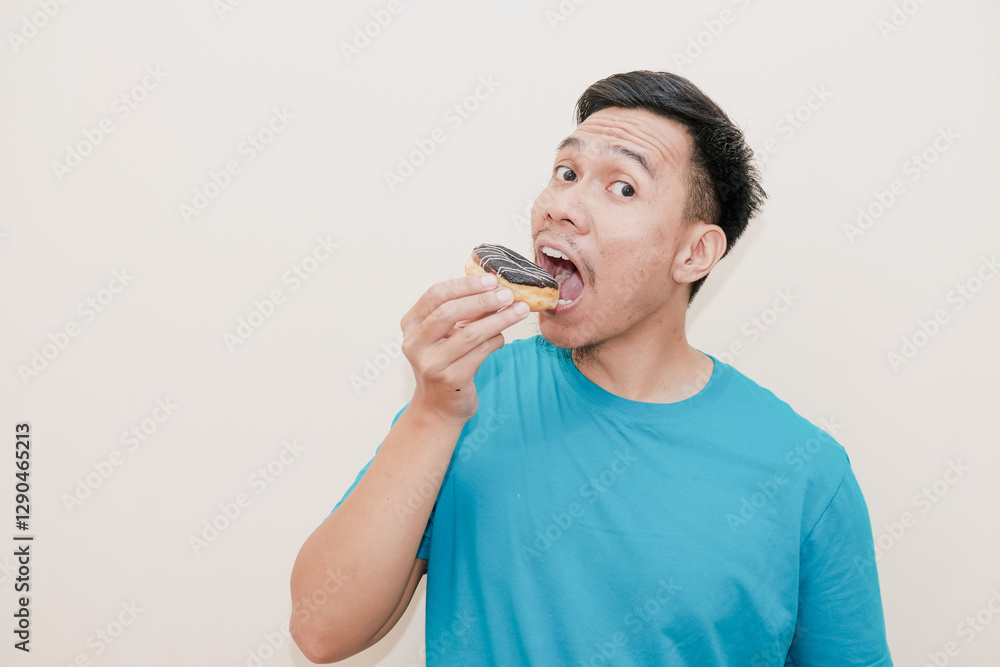 A funny Southeast Asian man with holding chocolate donut and eating tasty then with open mouth, in white background, delicious sweets and pastry concept A funny Southeast Asian man with holding chocolate donut and eating tasty then with open mouth, in white background, delicious sweets and pastry concept