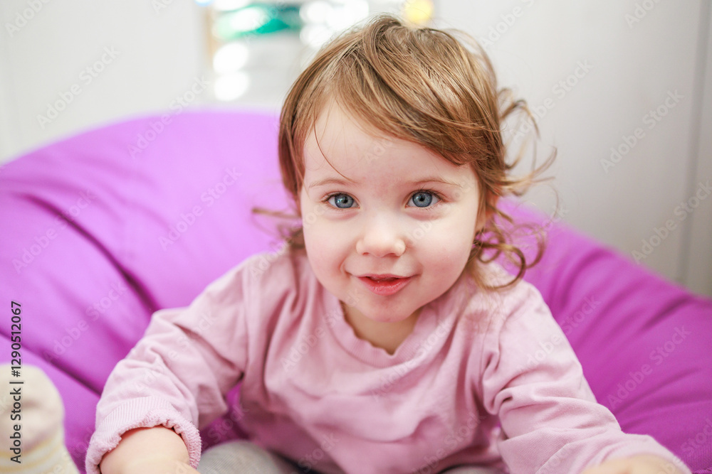A young girl with light brown hair and blue eyes is smiling. She is seated on a purple bean bag chair, creating a playful atmosphere. The background is softly out of focus A young girl with light brown hair and blue eyes is smiling. She is seated on a purple bean bag chair, creating a playful atmosphere. The background is softly out of focus