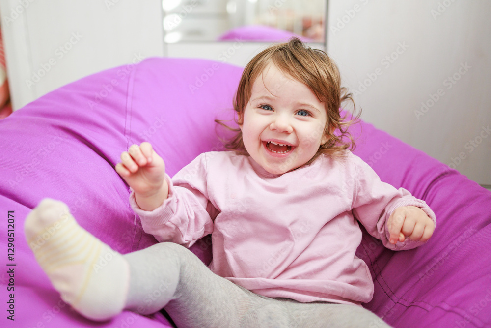 Fototapeta premium A young girl with light brown hair and blue eyes is smiling. She is seated on a purple bean bag chair, creating a playful atmosphere. The background is softly out of focus