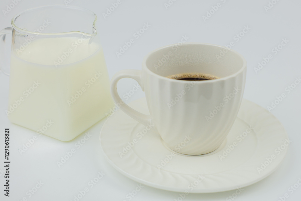 Close-up of a white mug with black coffee standing on a saucer, next to it is a jug of milk on a white background. The concept of invigorating drinks, coffee break, coffee with milk. High quality Close-up of a white mug with black coffee standing on a saucer, next to it is a jug of milk on a white background. The concept of invigorating drinks, coffee break, coffee with milk. High quality