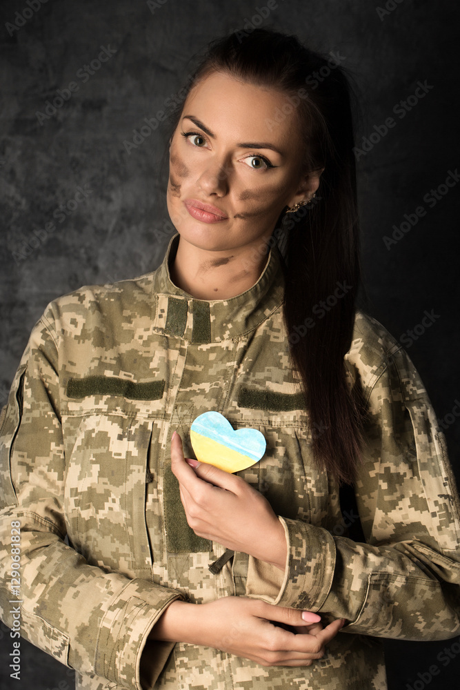 Beautiful military woman in military uniform with camouflage makeup on her face holds a heart-shaped Ukrainian flag in her hands