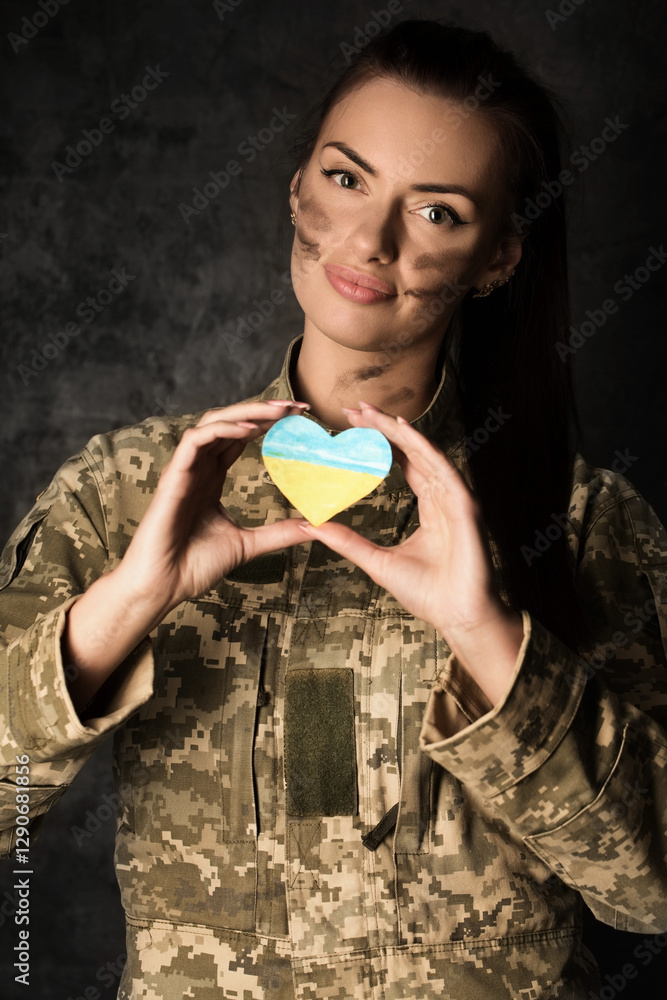 Military woman in military uniform with camouflage makeup on her face holds a heart-shaped Ukrainian flag in her hands