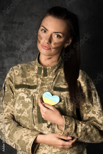 Obraz na płótnie Beautiful military woman in military uniform with camouflage makeup on her face holds a heart-shaped Ukrainian flag in her hands w salonie