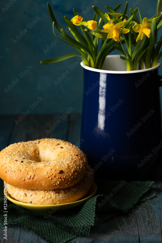 Still life in rustic style bagels sprinkled with seeds near a blue metal jug with yellow daffodils