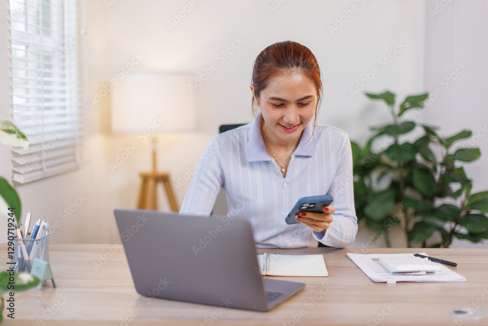 Serious charming asian woman using smartphone while working with laptop at home workplace.
Serious charming asian woman using smartphone while working with laptop at home workplace.