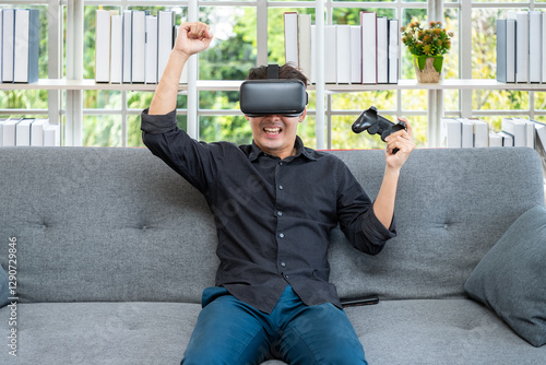 Asian man celebrating gaming victory while playing virtual reality VR game with headset and controller on sofa at home photo