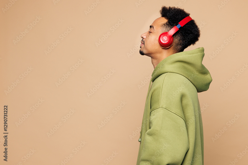 Φωτογραφία Side view young man of African American ethnicity he wear green hoodie casual clothes listen to music in headphones isolated on plain pastel light beige background studio portrait