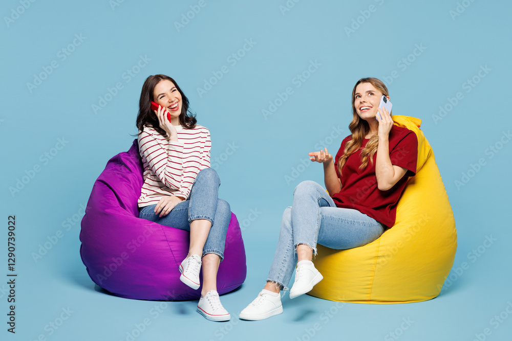 Φωτογραφία Full body young couple two friends women wearing red casual clothes together sit in bag chair talk speak on mobile cell phone isolated on pastel plain light blue background studio
