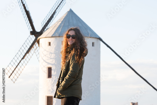Una mujer con gafas de sol y chaqueta verde posa junto a un tradicional molino de viento blanco en un paisaje rural. La luz cálida resalta la escena, evocando la belleza y la historia de Castilla