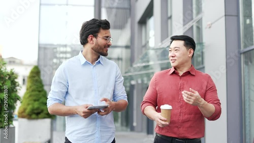 Two happy smiling businessmen engaging in discussion while walking outside modern office. One holds a tablet, another enjoys coffee. Teamwork and friendship among professionals in an urban setting