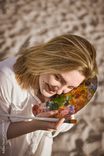 Cuadro en lienzo Young woman smiles serenely on sandy beach, gently holding colorful artist palette against her cheek