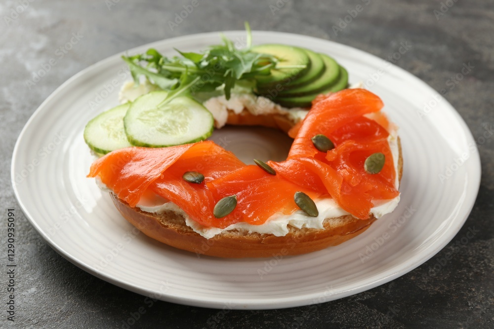 Delicious bagel with salmon, cream cheese, cucumber, avocado and pumpkin seeds on grey table, closeup Delicious bagel with salmon, cream cheese, cucumber, avocado and pumpkin seeds on grey table, closeup