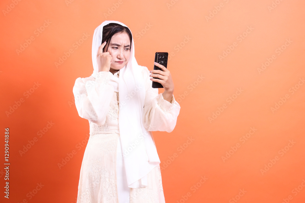 Confused Muslim Woman Staring at Smartphone: Depicting uncertainty and contemplation in modern communication, isolated on a orange background.