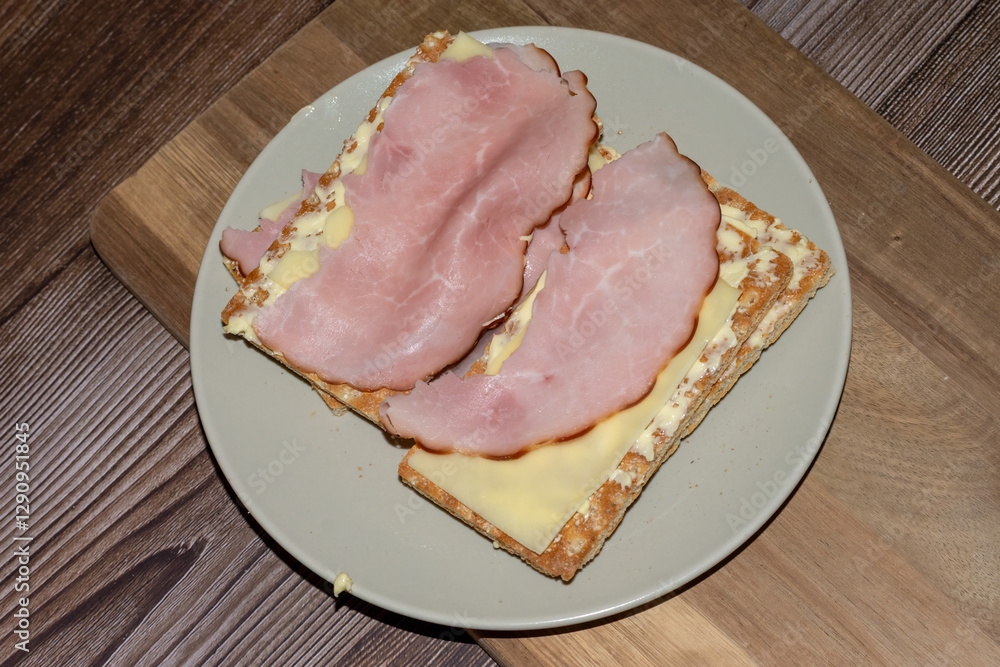 A simple open-faced sandwich features slices of ham and cheese on crisp bread, placed on a round plate. The setting shows a wooden table surface, creating a cozy atmosphere A simple open-faced sandwich features slices of ham and cheese on crisp bread, placed on a round plate. The setting shows a wooden table surface, creating a cozy atmosphere