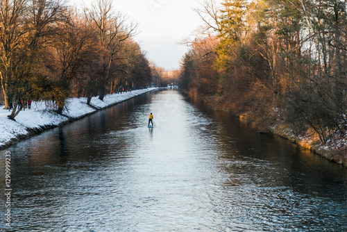 View of serene Isar river with a paddleboarder surrounded by snow-covered trees, Munich, Germany.