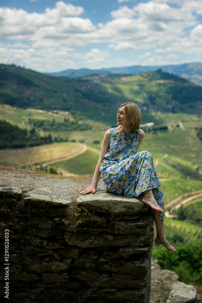 A woman in a light dress sits gracefully on a stone wall of a vineyard in the Douro Valley, Portugal.