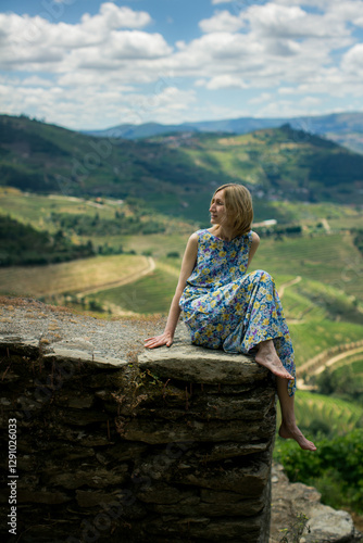 Obraz na płótnie A woman in a light dress sits gracefully on a stone wall of a vineyard in the Douro Valley, Portugal. w salonie