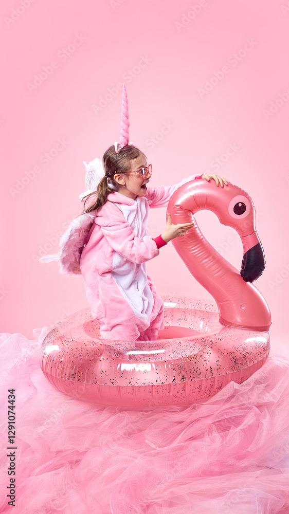 Obraz premium Girl in pink unicorn onesie excitedly playing with inflatable pink flamingo against pink background. Summertime vacation vibes. Concept of childhood, fantasy, dreams, imagination