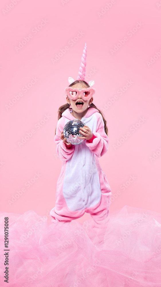 Obraz premium Happy girl in pink unicorn onesie laughing, wearing oversized pink glasses and holding disco ball against pink celebratory background. Concept of childhood, fantasy, dreams, imagination, party