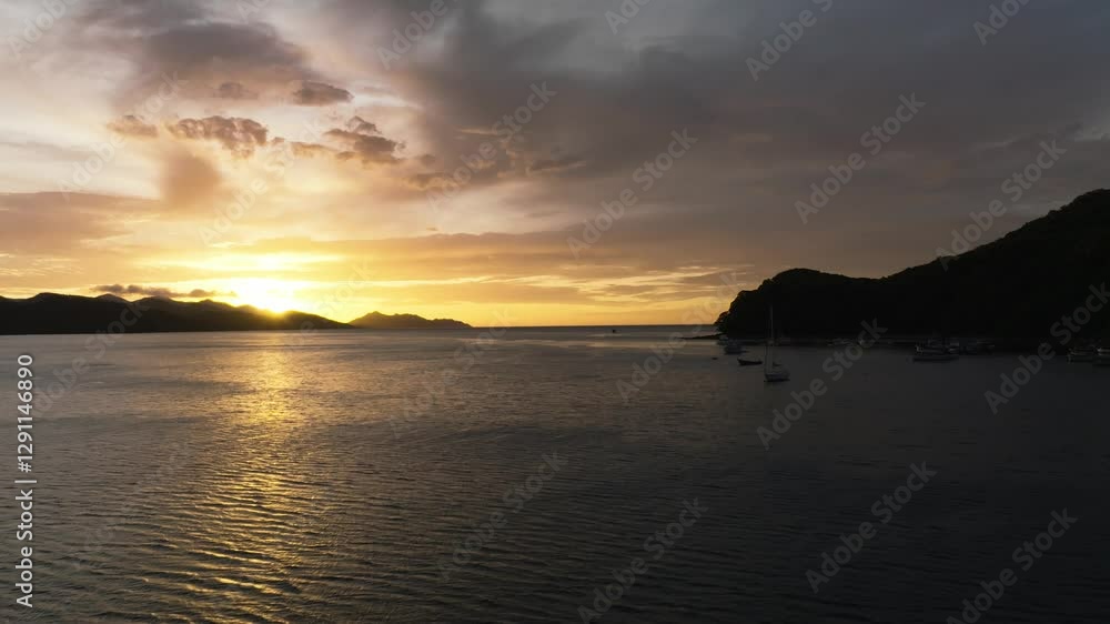 Aerial reverse tracking shot over Junquillal Bay, Costa Rica, at sunset. Golden light pierces clouds above silhouetted mountains, while anchored boats dot the shimmering waters. Aerial reverse tracking shot over Junquillal Bay, Costa Rica, at sunset. Golden light pierces clouds above silhouetted mountains, while anchored boats dot the shimmering waters.