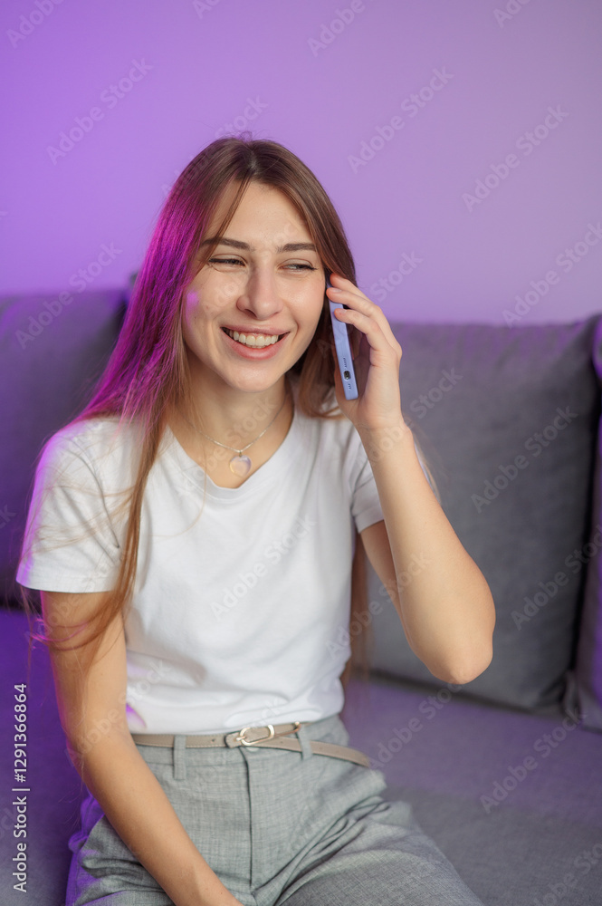 Cheerful smiling young woman is having a pleasant phone conversation, enjoying a moment of connection and laughter in the comfort of her home purple light Cheerful smiling young woman is having a pleasant phone conversation, enjoying a moment of connection and laughter in the comfort of her home purple light