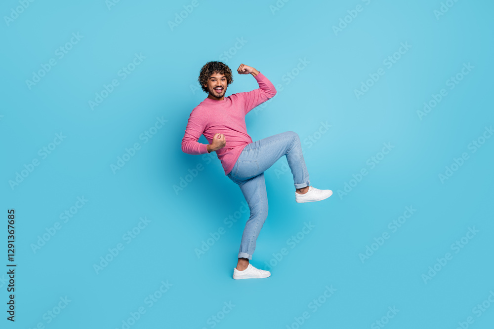 Fototapeta premium Happy stylish young man wearing pink pullover and denim jeans playfully posing on a blue background with a cheerful expression