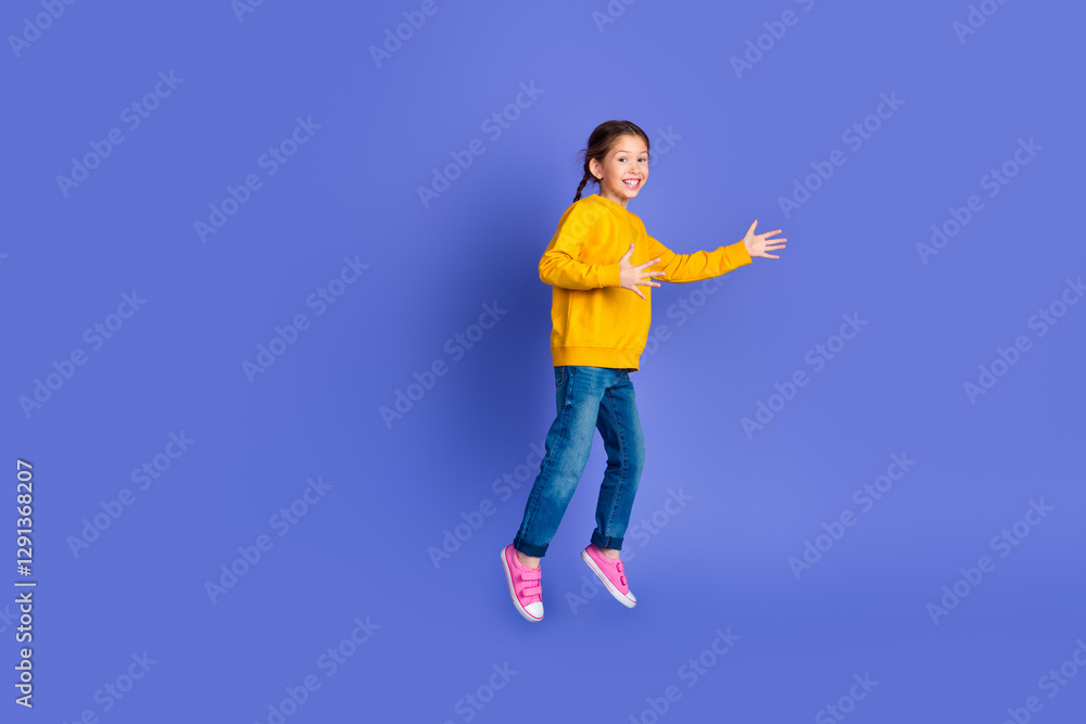 Joyful young girl wearing a yellow sweatshirt and jeans jumping on a vibrant purple background with a happy expression Joyful young girl wearing a yellow sweatshirt and jeans jumping on a vibrant purple background with a happy expression