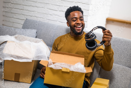 Cuadro en lienzo Happy young African-American man smiling and opening delivered package with headphones while sitting on sofa at his home