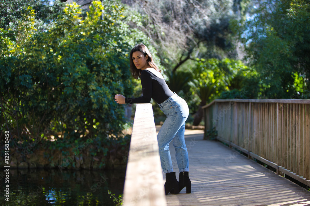 brunette woman, young and beautiful dressed in casual clothes leaning on the railing of a wooden bridge. The woman makes different body postures. In the background green vegetation. brunette woman, young and beautiful dressed in casual clothes leaning on the railing of a wooden bridge. The woman makes different body postures. In the background green vegetation.