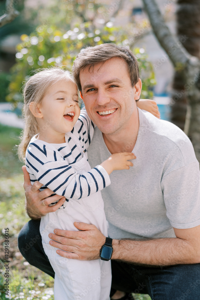 Little laughing girl hugs the neck of her smiling dad squatting in the garden Little laughing girl hugs the neck of her smiling dad squatting in the garden