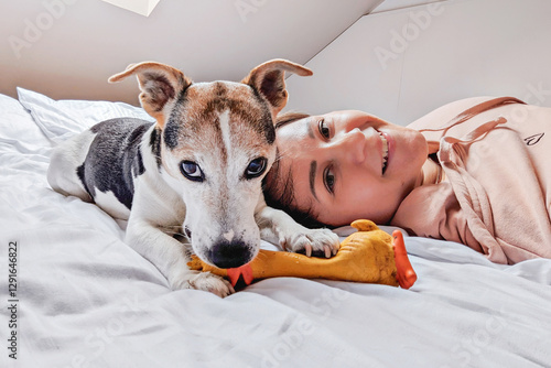 Wallpaper Mural Smiling woman lies on white bed next to her Jack Russell Terrier, who is chewing rubber toy, capturing warm and affectionate moment of companionship. Woman taking selfie with dog Torontodigital.ca