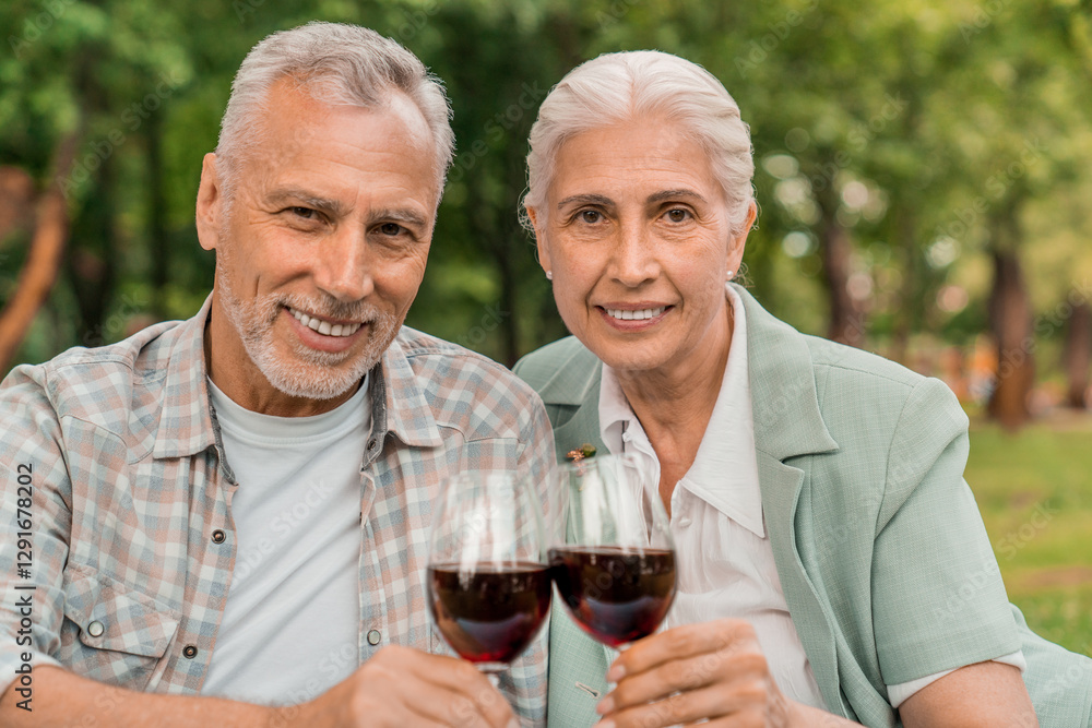 Senior Caucasian elderly couple happily embracing looking at camera and drinking red wine together during a picnic in the park outdoors. Happy retirement life concept Senior Caucasian elderly couple happily embracing looking at camera and drinking red wine together during a picnic in the park outdoors. Happy retirement life concept