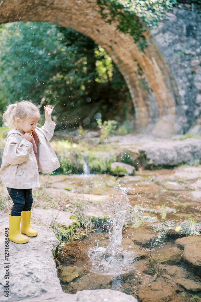 Little girl stands in splashes of water from a lake on a paved shore near a bridge in the park Little girl stands in splashes of water from a lake on a paved shore near a bridge in the park