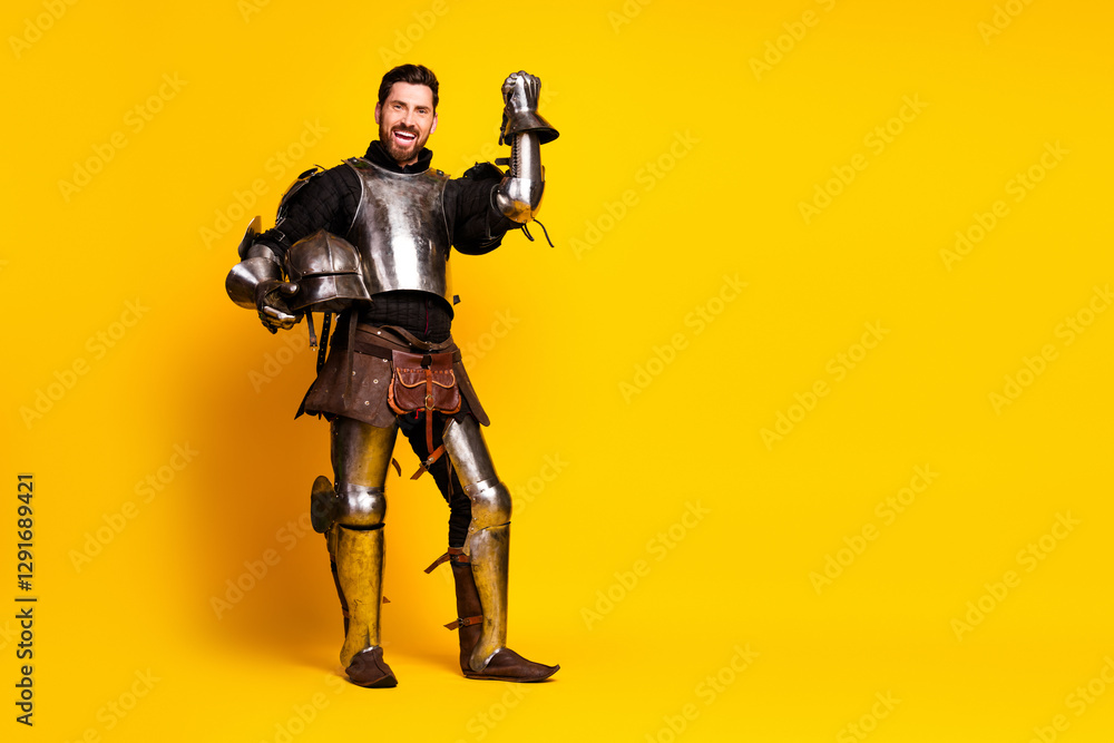 Young man in shiny medieval armor holding a helmet with a cheerful gesture on vibrant yellow background Young man in shiny medieval armor holding a helmet with a cheerful gesture on vibrant yellow background