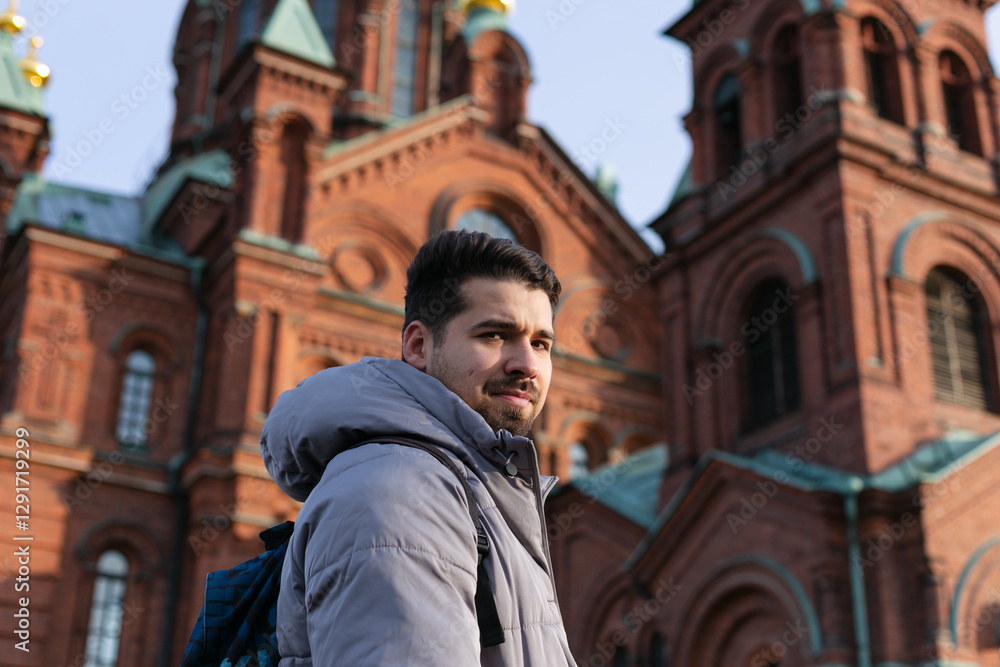 Naklejka premium Side view of a young adult traveler man in a grey puffer jacket standing in front of Helsinki’s Uspenski Cathedral. The red brick architecture and green domes contrast with the blue sky