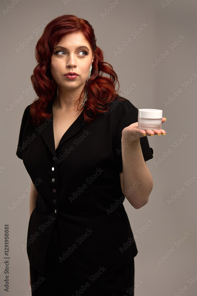 Elegant woman with wavy red hair and black dress holding a small jar and looking sideways, beauty and skincare concept