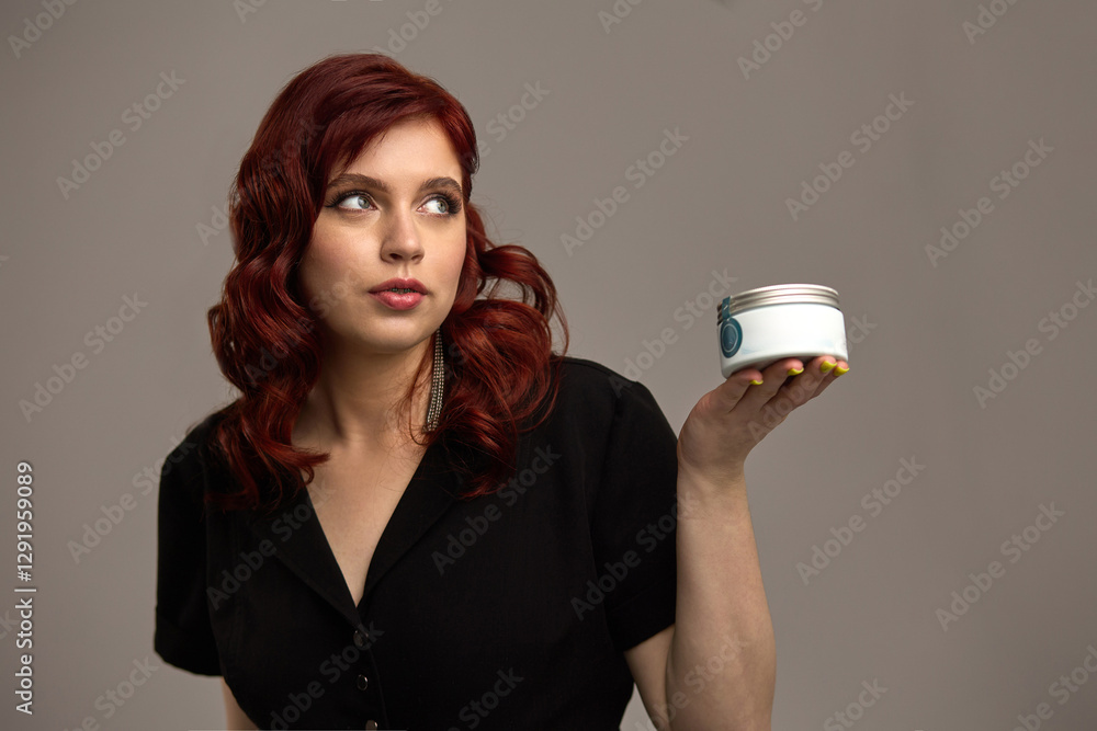 Stylish woman with red hair holding a cosmetic jar, thoughtful expression, beauty and skincare concept