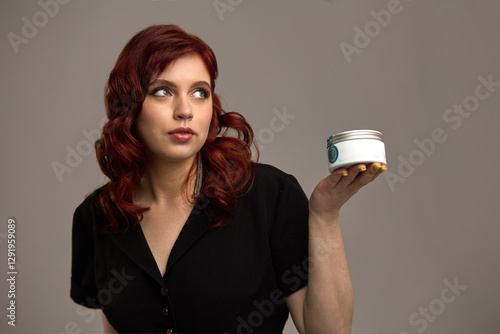 Obraz na płótnie Stylish woman with red hair holding a cosmetic jar, thoughtful expression, beauty and skincare concept w salonie