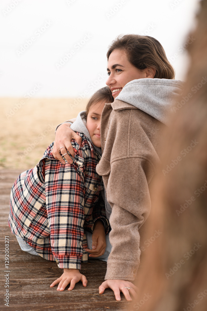 Fototapeta premium A mother and her daughter sit on the sand at the beach, laughing and hugging each other, enjoying their time together despite the chilly weather.