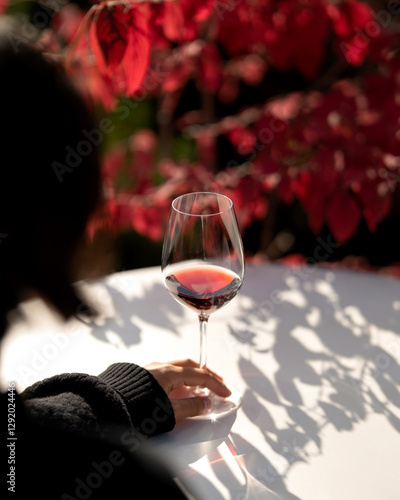 Burgundy wineglass with red wine in female hand on table. Ruby wine in bordeaux glass. Wine tasting. Aperitif. Alcohol beverage. Drink concept. Warm autumn atmosphere. Dinner in restaurant.