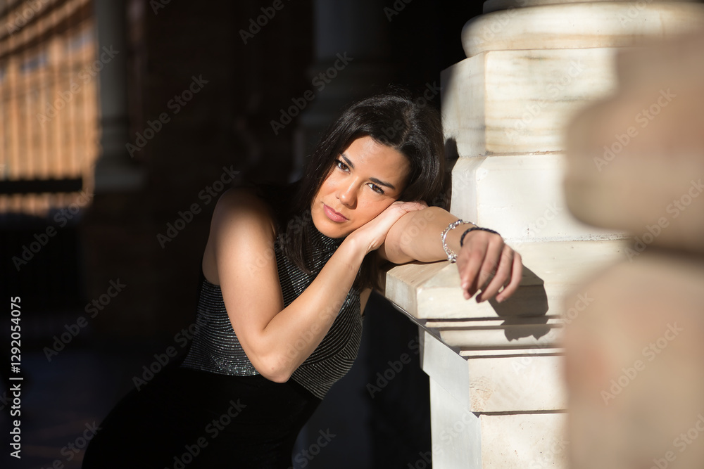 Portrait of young, beautiful, brunette woman, wearing a fancy black dress, looking at the camera, sweet and tender, leaning on a marble column, receiving the sun's rays. Concept beauty, femininity. Portrait of young, beautiful, brunette woman, wearing a fancy black dress, looking at the camera, sweet and tender, leaning on a marble column, receiving the sun's rays. Concept beauty, femininity.