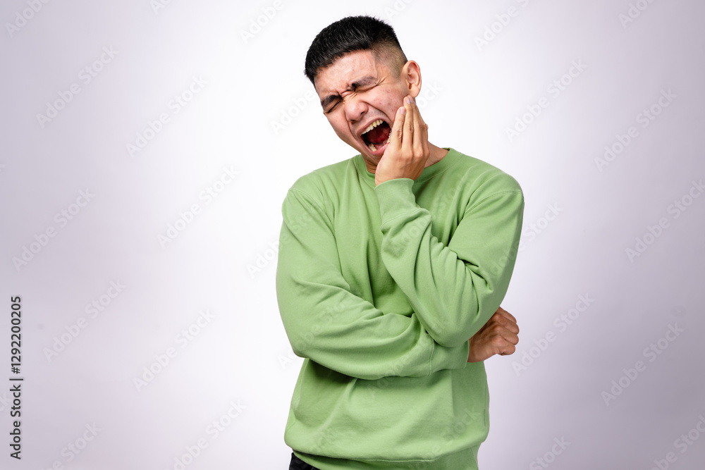 An Asian man in a green sweatshirt holds his cheek with one hand, eyes squinting and mouth open. His expression suggests a toothache or facial soreness against a white background An Asian man in a green sweatshirt holds his cheek with one hand, eyes squinting and mouth open. His expression suggests a toothache or facial soreness against a white background
