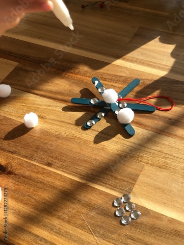 A child applies glue to create a snowflake-inspired Christmas ornament using popsicle sticks, pom poms, and shimmering gems on a wooden surface. photo