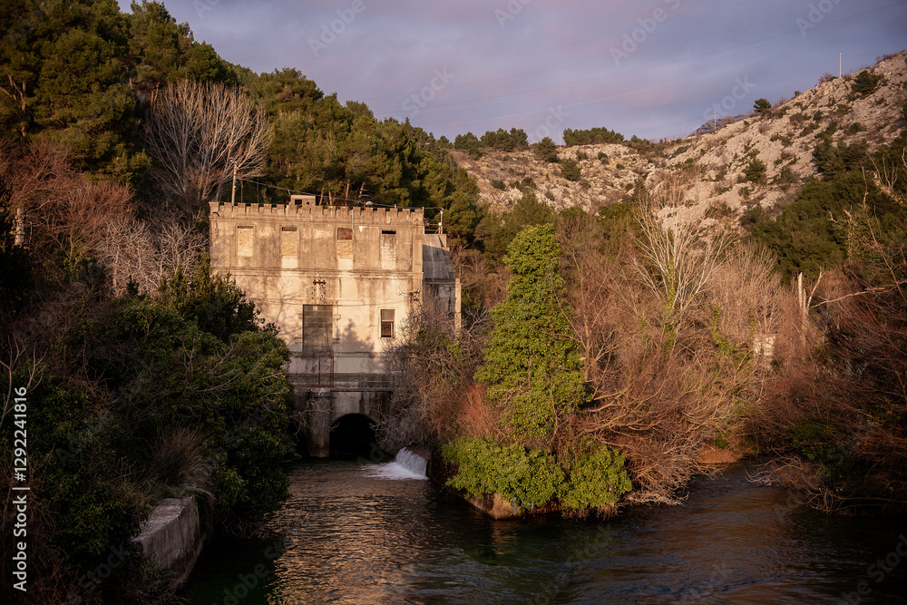 Fototapeta premium An old, abandoned building with a castle-like facade stands by a flowing river, surrounded by lush greenery and rocky hills, bathed in warm sunlight. Solin Croatia