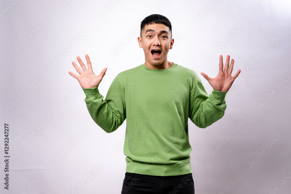 An excited Asian man in a green sweatshirt raises both hands with wide-open palms and a joyful expression. He stands against a plain white background, conveying surprise, excitement An excited Asian man in a green sweatshirt raises both hands with wide-open palms and a joyful expression. He stands against a plain white background, conveying surprise, excitement