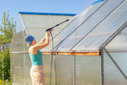 A woman cleaning a greenhouse roof with a pressure washer under bright sunlight. The image captures home farming and sustainable agriculture practices.