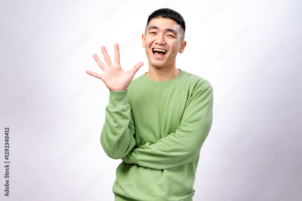 An Asian man in a green sweatshirt is smiling while holding up five fingers on his right hand. His left arm is crossed, and he stands against a white background, conveying a friendly An Asian man in a green sweatshirt is smiling while holding up five fingers on his right hand. His left arm is crossed, and he stands against a white background, conveying a friendly