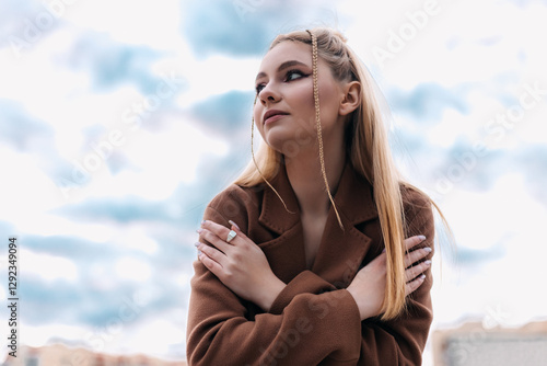 Fashionable young woman with braided hair wearing a brown coat, hugging herself and looking up at a cloudy sky, enjoying a peaceful moment outdoors