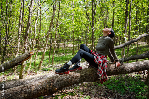 Young woman hiker is enjoying the peace of nature while sitting on a fallen tree trunk in a vibrant green forest. Taking a moment to breathe in the fresh air and appreciate the beauty surrounding her